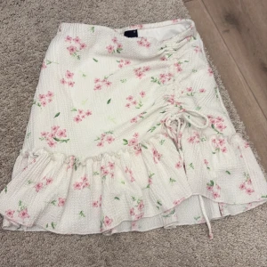 Vit blommig kortkjol från Gina Tricot - Supersöt vit kortkjol från Gina Tricot med rosa blommor och gröna blad. Kjolen har volangkant nedtill och snörning på sidan för extra detalj. Materialet är lätt och känns som bomull, perfekt för varma dagar. Passformen är normal och kjolen har en feminin vibe.