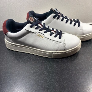 Vita Colmar sneakers med röd detalj - Snygga vita sneakers från Colmar med marinblå snörning och röd mockadetalj på hälen. Skorna har en klassisk rund tå, platt sula och Colmar-logga på sidan och bak. Perfekta för dig som gillar stilrena och sportiga skor med en twist.