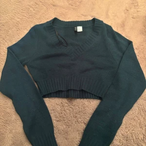 Mörkgrön croppad stickad tröja Divided - Snygg mörkgrön croppad stickad tröja från Divided med v-ringad hals och breda ribbade muddar. Perfekt för en trendig look med sina långa ärmar och tighta passform.