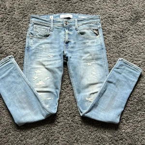 Replay ljusblå slitna jeans - Säljer ett par ljusblå jeans från Replay med snygga slitningar och klassisk femficksdesign. Jeansen har raka ben och normal passform, med läderpatch bak i midjan och dragkedjegylf. Perfekta för en avslappnad streetstil.