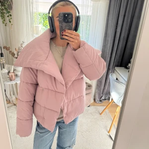 Ljusrosa pufferjacka med stor krage - Säljer en ljusrosa pufferjacka med oversized passform och stor, bred krage. Jackan har knäppning framtill och är quiltad för extra volym. Perfekt för dig som vill ha en trendig och mysig look under kyliga dagar.
