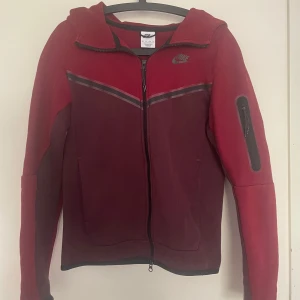 Nike tech  fleece - Snygg och bekväm Nike-hoodie med hel dragkedja. Modellen har en tvåfärgad design i rött och vinrött med svart Nike-logga på bröstet och diskret randdetalj över bröstet. 	•	Stor huva med bra passform 	•	Två stora framfickor 	•	Dragkedja som kan öppnas både uppifrån och nerifrån 	•	Muddar i svart kantband vid ärmslut och nederkant 	•	Perfekt för träning, fritid eller vardagsbruk  Plagg i galant skick, endast använt ett fåtal gånger. Broschyren säger xs men passar mer S samt M