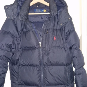 Mörkblå dunjacka Polo Ralph Lauren - Snygg mörkblå dunjacka från Polo Ralph Lauren med klassisk röd logga på bröstet. Jackan har dragkedja framtill, hög krage och praktiska fickor. Perfekt för kalla dagar och riktigt stilren att bära till det mesta.