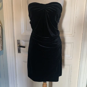 Svart axelbandslös sammetsklänning - Snygg svart axelbandslös klänning i mjuk sammet med rynkade detaljer framtill och längs sidorna. Klänningen har en figurnära passform upptill och faller rakt ner till knäna. Perfekt för dig som vill ha en stilren och elegant look.