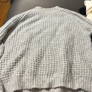 Grå stickad tröja med struktur - Säljer en grå stickad tröja med snyggt strukturerat mönster och ribbade muddar. Perfekt för kyliga dagar och enkel att matcha med jeans eller kjol. Rund halsringning och långärmad modell.