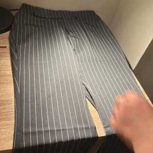 Svarta randiga bootcut kostymbyxor - Snygga svarta kostymbyxor med vita vertikala ränder och bootcut-ben. Byxorna har klassisk passform med medelhög midja och är perfekta för en stilren look. Materialet känns mjukt och följsamt, och de har hällor för bälte.