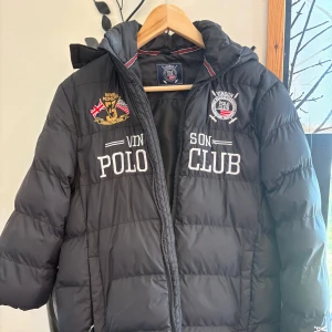 Svart pufferjacka från Vinson Polo Club 160 storlek  - Svart pufferjacka från Vinson Polo Club med broderade märken och loggor på bröstet. Jackan har huva, dragkedja framtill och quiltad design. Perfekt för kyliga dagar och ger en sportig look.