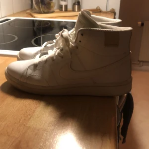 Vita höga sneakers från Nike - Säljer ett par klassiska vita höga sneakers från Nike med snörning och rund tå. Skorna är i skinn och har en stilren look med diskret Nike-logga på sidan och bak. Perfekta för dig som gillar en clean och tidlös stil.