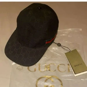 Gucci Keps - Svart keps från Gucci med klassiskt GG-monogram över hela kepsen och ikoniskt grönt och rött band på sidan. Justerbar rem baktill i skinn. Tillverkad i Italien och levereras med originalpåse och tagg. Perfekt för dig som vill ha en lyxig och stilren accessoar.