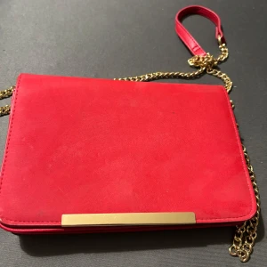 Röd mocka kuvertväska med guldkedja - Snygg röd kuvertväska i mocka med guldfärgad kedja och stilren metallist framtill. Perfekt för dig som vill sticka ut med en färgklick och gillar trendiga accessoarer. Enkel att bära över axeln tack vare kedjan.