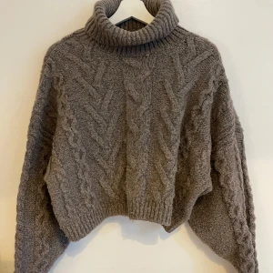 Brun/ beige polotröja i ullmix från H&M - Mysig brun polotröja från H&M i en skön ullblandning. Tröjan har grovstickat flätmönster, oversized passform och höga muddar. Perfekt för kalla dagar när du vill vara både varm och snygg. XL storlek men är croppad. 