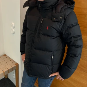 Svart pufferjacka från Polo Ralph Lauren - Svart pufferjacka från Polo Ralph Lauren med röd logga på bröstet. Jackan har huva, flera dragkedjeförsedda fickor och quiltad design. Perfekt för kalla dagar och har en riktigt snygg streetstil. Materialet är syntet och jackan är långärmad. 