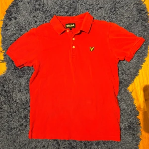 Röd pikétröja från Lyle & Scott - Röd pikétröja från Lyle & Scott med klassisk krage och knappar framtill. Tröjan har korta ärmar och ikonisk gul broderad örn på bröstet. Tillverkad i bomull för en skön och luftig känsla. Perfekt för en stilren och sportig look.