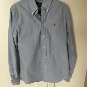 Blå skjorta från Polo Ralph Lauren(pris kan diskuteras) - Klassisk ljusblå skjorta från Polo Ralph Lauren med broderad logga på bröstet. Skjortan har lång ärm, knappar framtill och button-down krage. Modellen är custom fit och tillverkad i bomull för en skön känsla.