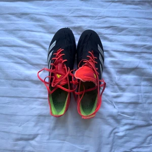 Adidas Predator 42 - Säljer ett par Adidas Predator fotbollsskor i svart med röda och vita detaljer. Skorna har snörning, mönstrad ovansida och dobbar för gräsplan. Insidan är grön och sulan har en cool röd-vit design. Har bara en skavank på framsidan av ena skorn. Perfekta för dig som vill sticka ut på planen. storlek 42.