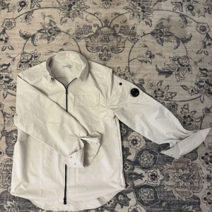 Cp company overshirt  - Säljer en stilren vit overshirt från C.P. Company med svart linsdetalj på ärmen och bröstficka. Skjortan har dragkedja framtill och klassisk krage. Perfekt för dig som gillar minimalistisk och modern streetwear. Anständig 2 gånger och nästa helt ny