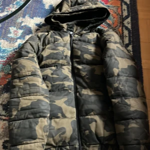 Camouflage pufferjacka från Zara - Säljer en fet pufferjacka från Zara med camouflage-mönster i gröna och beige toner. Jackan har huva, dragkedja framtill och långa ärmar med muddar. Perfekt för kalla dagar när du vill sticka ut lite extra.