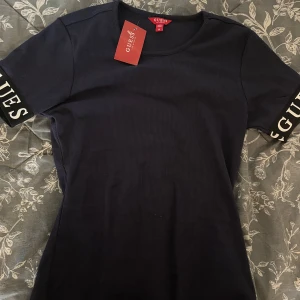 Svart ribbad topp från Guess - Säljer en svart ribbad topp från Guess med korta ärmar. Ärmarna har breda muddar med vit GUESS-logga tryckt på. Toppen har rund halsringning och är i stretchigt material som sitter snyggt på kroppen.