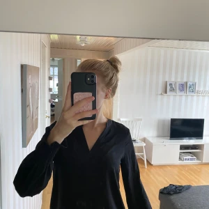 Svart v-ringad blus från Vero Moda - Svart blus från Vero Moda i storlek XS med v-ringad hals och långa ärmar. Ärmsluten har snygg smockdetalj som ger en ballongliknande effekt. Tillverkad i mjuk bomull, perfekt för en clean och stilren look.