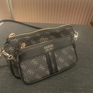 Axelväska guess - Snygg svart axelväska från Guess med klassiskt logomönster i grått och guldfärgade detaljer. Väskan har flera fack med dragkedja och justerbar axelrem. Den är använd, men fint skick. De ända synliga som syns är vid metallen där guld färgen missfärgats lite men de syns inte överdrivet mycket. 