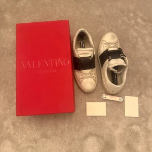 Valentino Garavani vita sneakers - Snygga vita sneakers från Valentino Garavani med bred svart rem över vristen och klassisk logga på plösen. Skorna har rund tå, snörning och platt sula. Kommer med originalkartong och extra skosnören. Perfekta för dig som gillar exklusiv streetstyle.