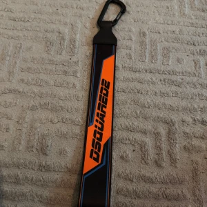 Svart och orange Dsquared2 keychain - Säljer ett snyggt keychain från Dsquared2 med coolt grafiskt tryck i orange och svart, samt blå detaljer. Bältet har en modern design med karbinhake i svart plast och bred rem. Perfekt för dig som vill sticka ut med en unik accessoar.