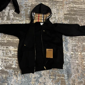 Svart Burberry hoodie med dragkedja XL - Svart hoodie från Burberry i storlek XL med klassiskt rutigt foder i huvan. Tröjan har dragkedja, snörning i huvan och fickor framtill. Guldiga detaljer på snörningen och dragkedjan ger en lyxig känsla. Perfekt för dig som vill ha en stilren och exklusiv look.