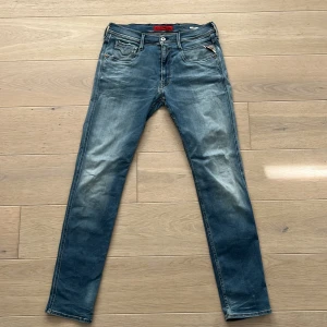 Replay Anbass 31 - Snygga Replay Anbass jeans i blå tvätt med slitningar och klassiska fem fickor. Slim fit-modell med smalare ben och låg midja. Jeansen har detaljer som logopatch bak och dragkedja i gylfen. Perfekta för dig som gillar en modern och stilren look.