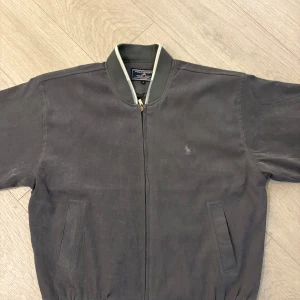  bomberjacka Polo Ralph Lauren - Snygg grå bomberjacka från Polo Sport Ralph Lauren med klassisk ribbad krage och dragkedja framtill. Jackan har två fickor på sidorna och diskret logga broderad på bröstet. Perfekt för dig som gillar stilren och sportig look. Jackan är producerad i 1990 talet, och är en jätte unik jacka, gjord av ett väldig skön material som är perfekt för hösten/vintern och jättestylig. Mycket Bra Skick! 