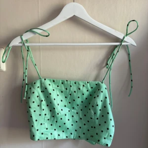 Mintgrön prickig topp från Zara - Superfin mintgrön topp från Zara med svarta prickar och smala axelband som knyts upptill. Croppad modell med lätt glansigt tyg och smockad baksida för extra komfort. Perfekt för dig som gillar färg och lekfulla detaljer!