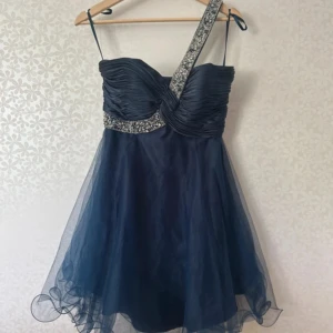 Marinblå kort klänning med paljetter - Säljer en marinblå kort klänning med ett lager av tunn tyll och snygga paljett-detaljer på midjan och axelbandet. Klänningen har ett oneshoulder-band och ett draperat liv som ger en lyxig känsla. Perfekt för dig som vill sticka ut med glittriga detaljer och cool design. 