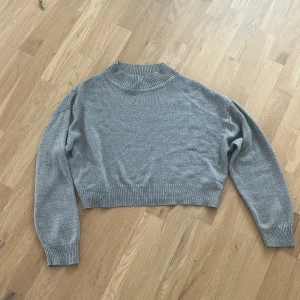 Grå croppad stickad tröja H&M Divided - Grå croppad stickad tröja från H&M Divided med ribbade muddar och högre hals. Perfekt till jeans eller kjol för en chill look. Långärmad och i mjukt material som passar till lager på lager under höst och vinter.