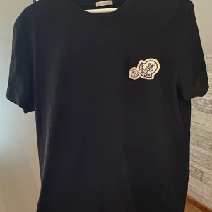 Svart Moncler t-shirt med logga - Snygg svart t-shirt från Moncler i bomull med klassisk rund hals och korta ärmar. På bröstet sitter en broderad Moncler-logga som ger en exklusiv touch. Perfekt för dig som gillar stilrena och trendiga plagg. Storlek L slim fit