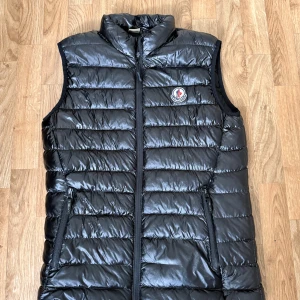 Svart dunväst från Moncler - Snygg svart dunväst från Moncler med glansig finish och klassisk logga på bröstet. Västen har quiltad design, två fickor med dragkedja och hög krage. Insidan har en röd, vit och blå randig detalj längs dragkedjan. Perfekt för lager-på-lager-stil. Lite sönder!!
