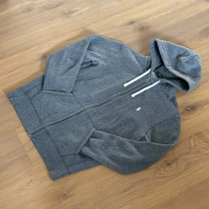 Grå hoodie från Polo Ralph Lauren - Mysig grå hoodie från Polo Ralph Lauren med vit logga broderad på bröstet. Tröjan har dragkedja framtill, huva med vita snören och två fickor. Perfekt för chill dagar och enkel att matcha med jeans eller joggers.