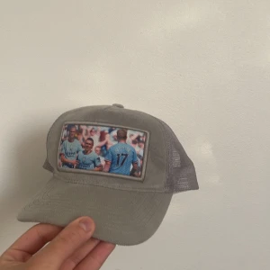 Beige meshkeps med fotbollstryck - Säljer en beige keps med mesh på sidorna och baksidan för extra ventilation. Framtill finns ett tryck med fotbollsspelare i ljusblå tröjor. Kepsen har böjd skärm och justerbar passform. Perfekt för dig som gillar sport och vill ha en unik accessoar.