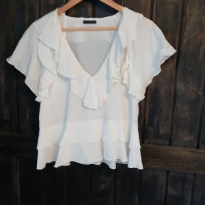 Vit volangblus från Zara - Supersöt vit blus från Zara med v-ringning och stora volanger längs halsringningen och ärmarna. Blusen är luftig och har korta, vida ärmar samt dubbla volanglager nertill. Perfekt för dig som gillar en romantisk och trendig look.