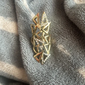 Guldig statementring med glittriga stenar - Säljer en unik och edgy guldig ring med geometriska trianglar och glittriga stenar. Ringen är stor och täcker mycket av fingret, perfekt för dig som gillar att sticka ut. Den har öppet band så storleken kan justeras. Ingen märkning syns.