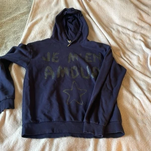 Mörkblå hoodie från Gina Tricot - Mörkblå hoodie från Gina Tricot med glittrig text 'JE M'EN AMOUR' och en stjärna framtill. Tröjan har huva, långa ärmar och ribbade muddar. Perfekt för dig som gillar en chill och snygg stil.