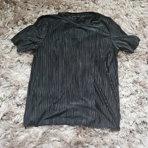 Svart plisserad topp från Mywear - Säljer en svart plisserad topp från Mywear i storlek M. Toppen har korta ärmar och är gjord i ett glansigt, lätt material som ger en snygg struktur. Perfekt för dig som gillar stilrena och trendiga plagg med det lilla extra.