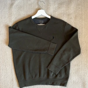 Mörkgrön sweatshirt från Polo Ralph Lauren - En stilren mörkgrön sweatshirt från Polo Ralph Lauren med rund halsringning och broderad logga på bröstet. Tröjan har långa ärmar och är tillverkad i mjuk bomullsmix, perfekt för lager-på-lager under hösten och vintern. Nypris ca 1200-1500kr.