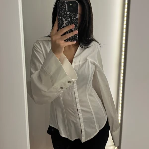 Corset skjorta - Säljer en vit skjorta med breda, långa ärmar och markerad midja. Skjortan har klassisk krage, knappar framtill och stilrena sömmar som ger en snygg siluett. 