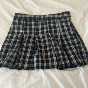 Svartvit rutig kjol från H&M Divided - Kort plisserad kjol från H&M Divided i svartvitt rutigt mönster. Kjolen har bred linning och klassiska veck som ger en snygg siluett. Perfekt för dig som gillar en preppy och trendig stil.