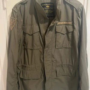 Grön overshirt från Alpha Industries - Säljer en militärinspirerad grön overshirt från Alpha Industries med patchar och flera fickor framtill. Jackan är tillverkad i bomull och polyester, har klassisk krage och knäppning framtill. Perfekt för dig som gillar utility-stil och streetwear.