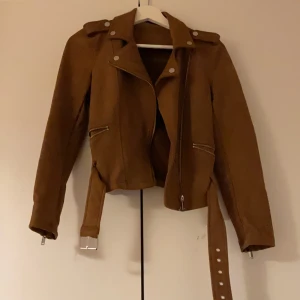 Brun mocka bikerjacka med bälte - Snygg brun bikerjacka i mocka med coola silverdetaljer, dragkedjor och bälte i midjan. Jackan har långa ärmar, axelklaffar och en croppad passform som ger en edgy vibe. Perfekt för dig som vill sticka ut med stilren look.