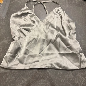 Silvrig omlottopp från NA-KD - Säljer en silvrig omlottopp från NA-KD i glansigt satinliknande material. Toppen har smala axelband som korsas i ryggen och en snygg omlottdesign framtill. Perfekt för dig som gillar stilrena och trendiga plagg med lite extra shine.