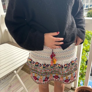 Vit broderad kortkjol med tofsar - Superfin vit kortkjol med broderade blommor i flera färger och spegeldekorationer. Kjolen har smock i midjan, volangkant och färgglada tofsar framtill. Perfekt för dig som gillar boho-stil och detaljerade plagg💐💐💐💐