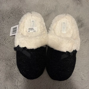 Svarta glittriga tofflor med rosett - Mysiga slip-in tofflor i svart med glittrig ovandel och fluffig vit insida. Dekorerade med en liten svart rosett på ovansidan. Perfekta för att hålla fötterna varma hemma och har en platt, vit sula för extra komfort. Aldrig använda! 