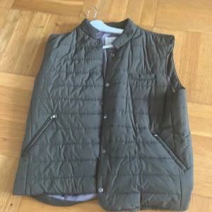 Quiltad dunväst från Four Hands - Stilren quiltad dunväst från Four Hands i mörkgrön färg. Västen har tryckknappar framtill, två snedställda fickor med dragkedja och en mjuk, ljusgrå insida. Perfekt för lager-på-lager och snygg till både hoodie och skjorta.