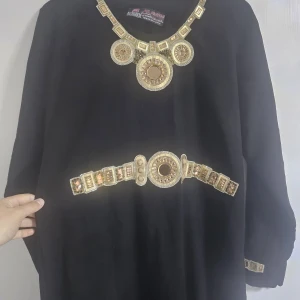 Svart långklänning med guldiga detaljer - Svart långklänning från Al-Madina med unika guldiga dekorationer vid halsringning, midja och ärmslut. Klänningen har lång ärm och ett elegant, löst fall. Materialet känns mjukt och har en lyxig look, perfekt för dig som gillar statement-plagg med bling.